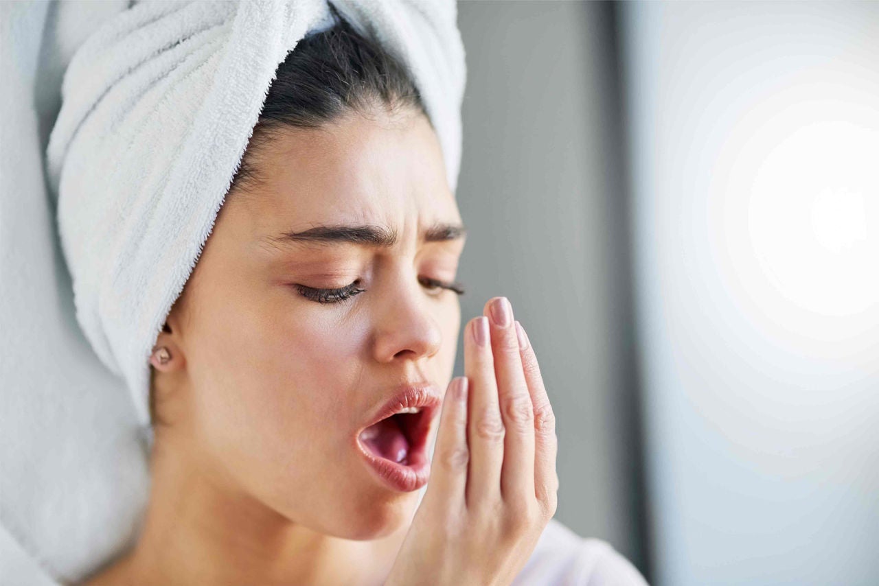 Mulher com toalha no cabelo e olhos fechados com expressão de desconforto, segurando a mão perto da boca, sentindo seu próprio hálito.