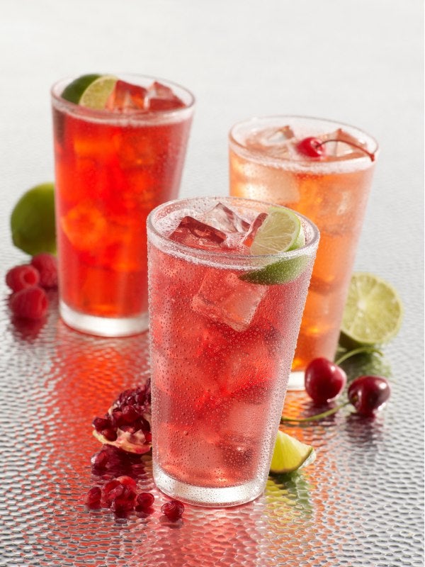 Três copos de soda italiana colorida com frutas como cereja, romã e limão, acompanhados de gelo.