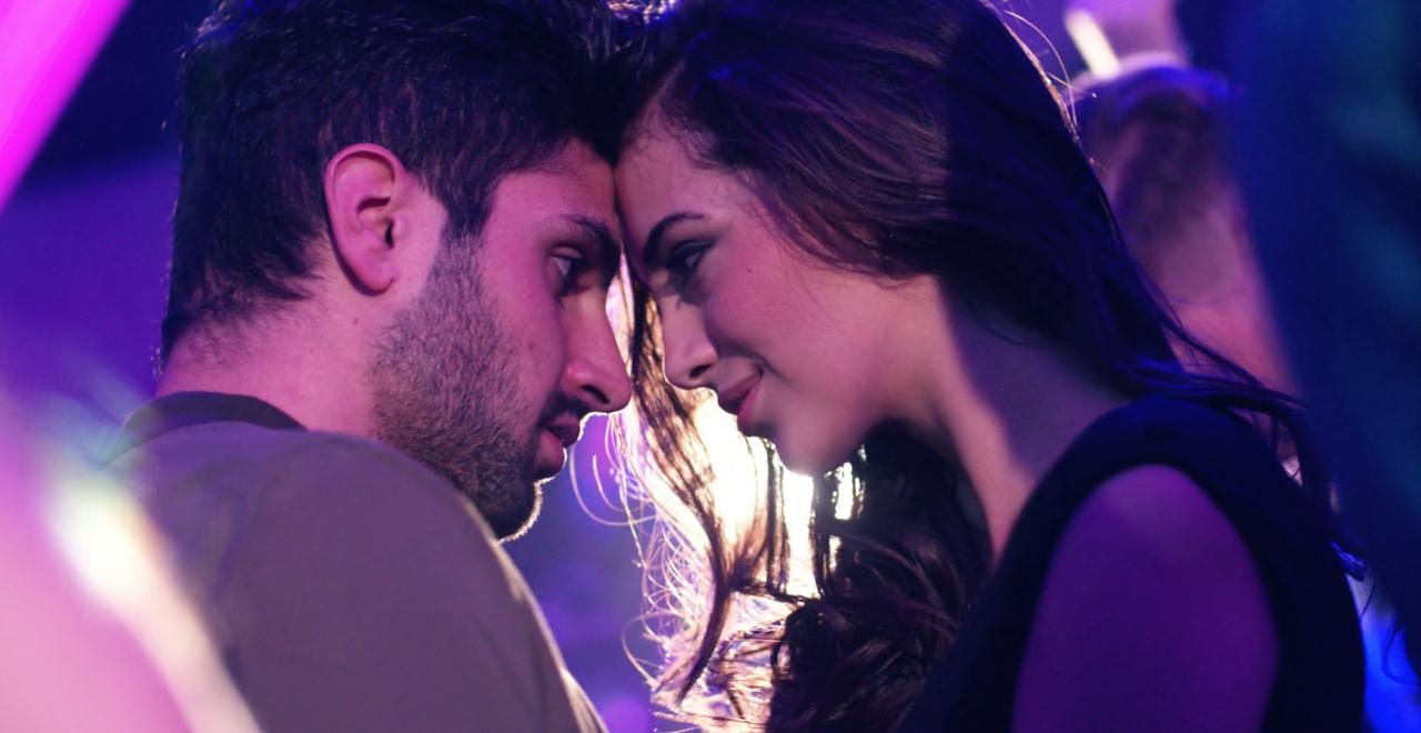Homem e mulher em clima de romance, com olhar intenso, prestes a dar um beijo de língua sob as luzes de uma festa. 