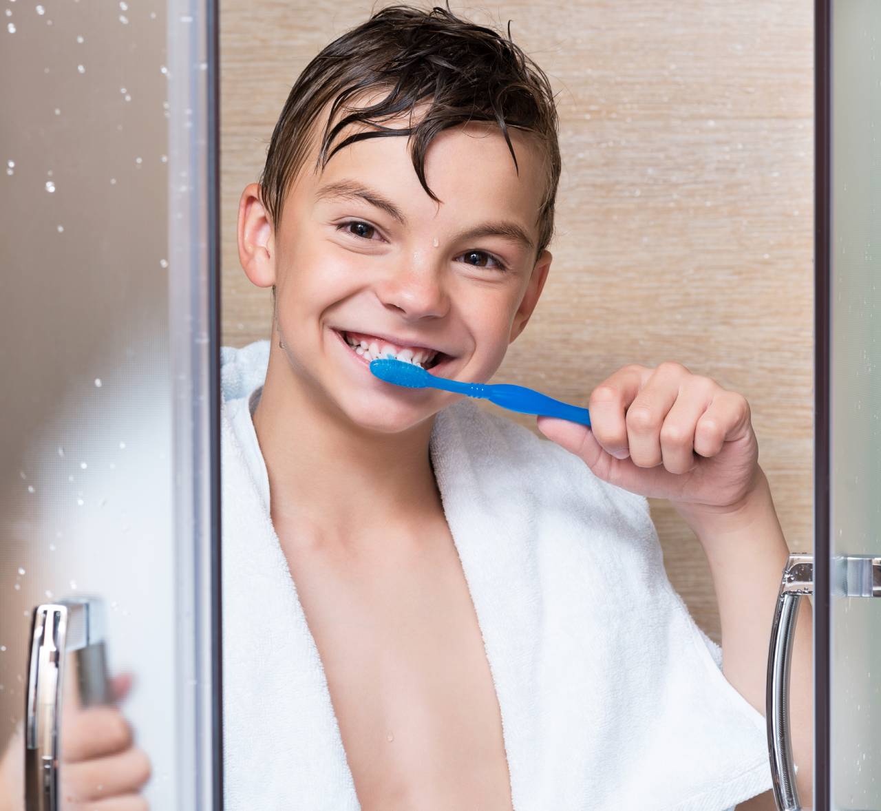 Menino sorridente com cabelo molhado e toalha branca nos ombros, mostrando o hábito de escovar dente no banho.