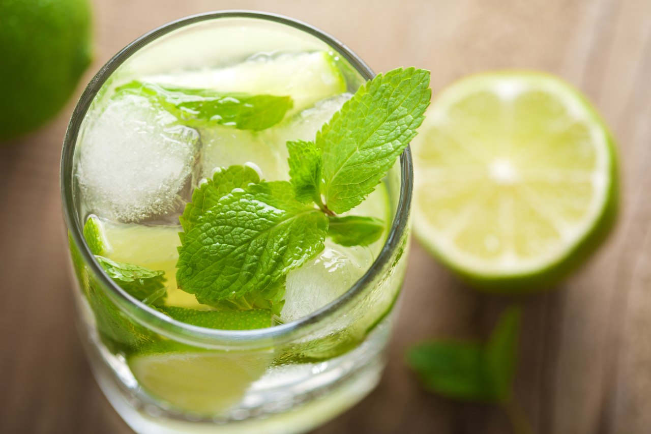 Copo de limonada suíça com gelo, folhas de hortelã e pedaços de limão, em uma mesa de madeira com meio limão ao fundo.