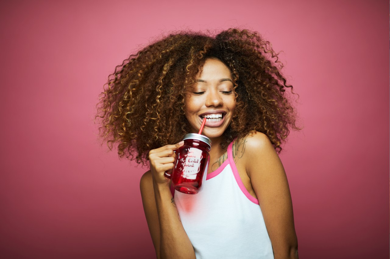 Mulher sorridente segura um copo com soda italiana vermelha, usando canudo, em frente a um fundo rosa vibrante. Mulher sorridente segura um copo com soda italiana vermelha, usando canudo, em frente a um fundo rosa vibrante.