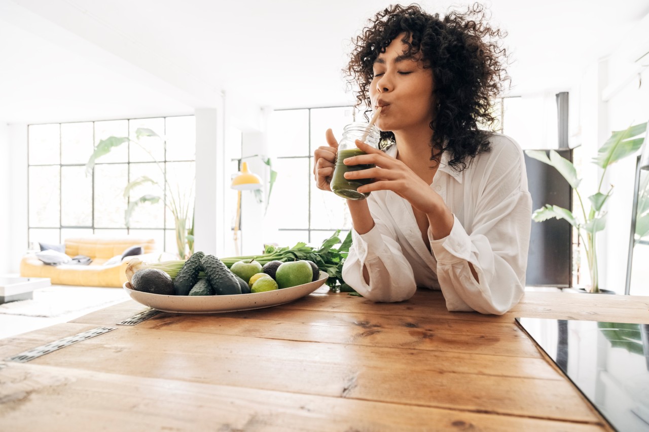 Mulher tomando suco verde na cozinha ao lado de gamela com alimentos naturais verdes.